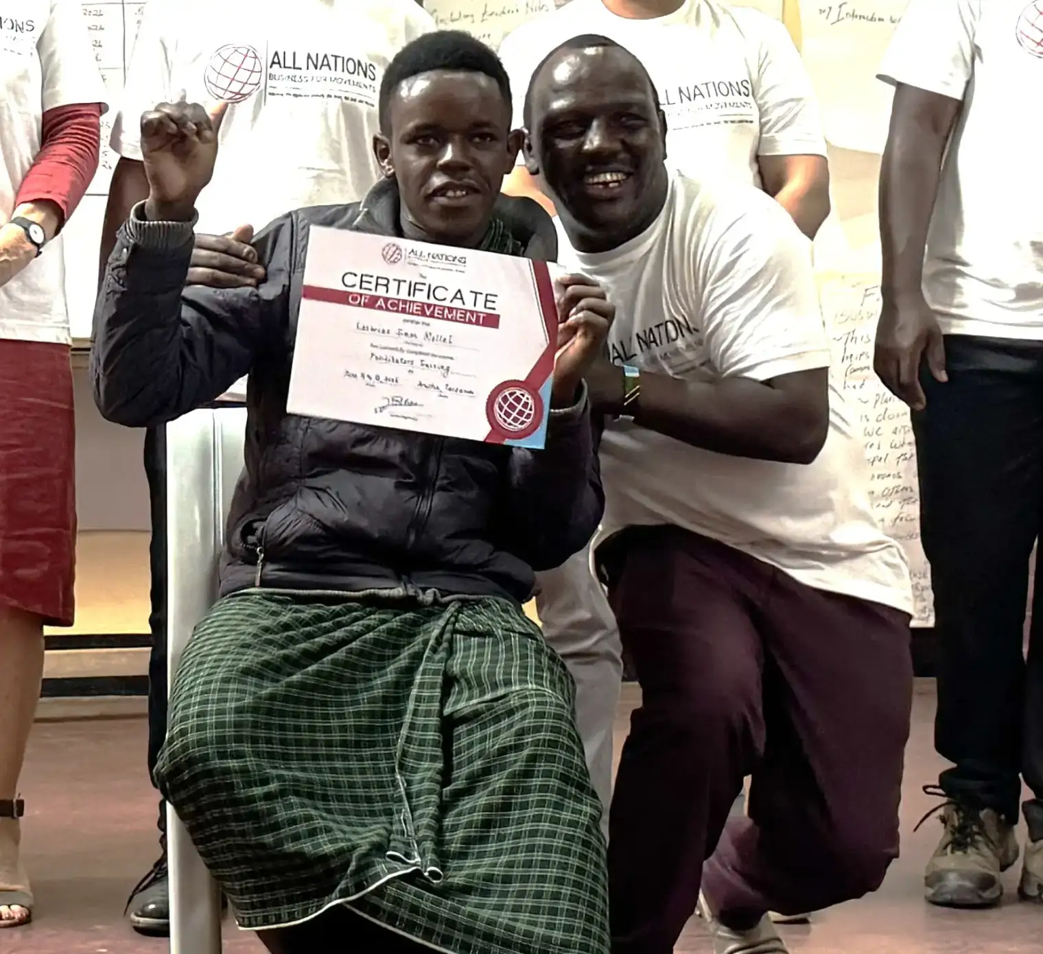 Loserian holding up his certificate of achievement from the Facilitators Training, next to a smiling man in an All Nations T-shirt