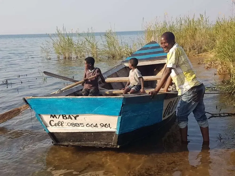 A man stands in shallow lake water pushing a blue and white wooden fishing boat with two boys sitting in it, Malawi.