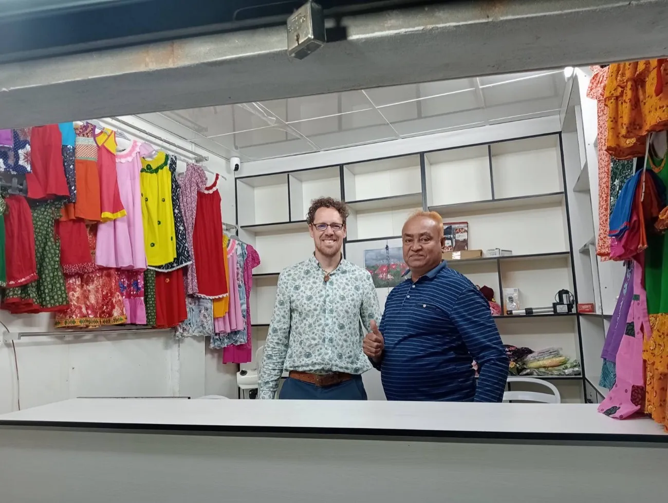 Two smiling men stand behind the counter of a clothing shop with brightly coloured dresses on the walls, one giving a thumbs up, Bangladesh.