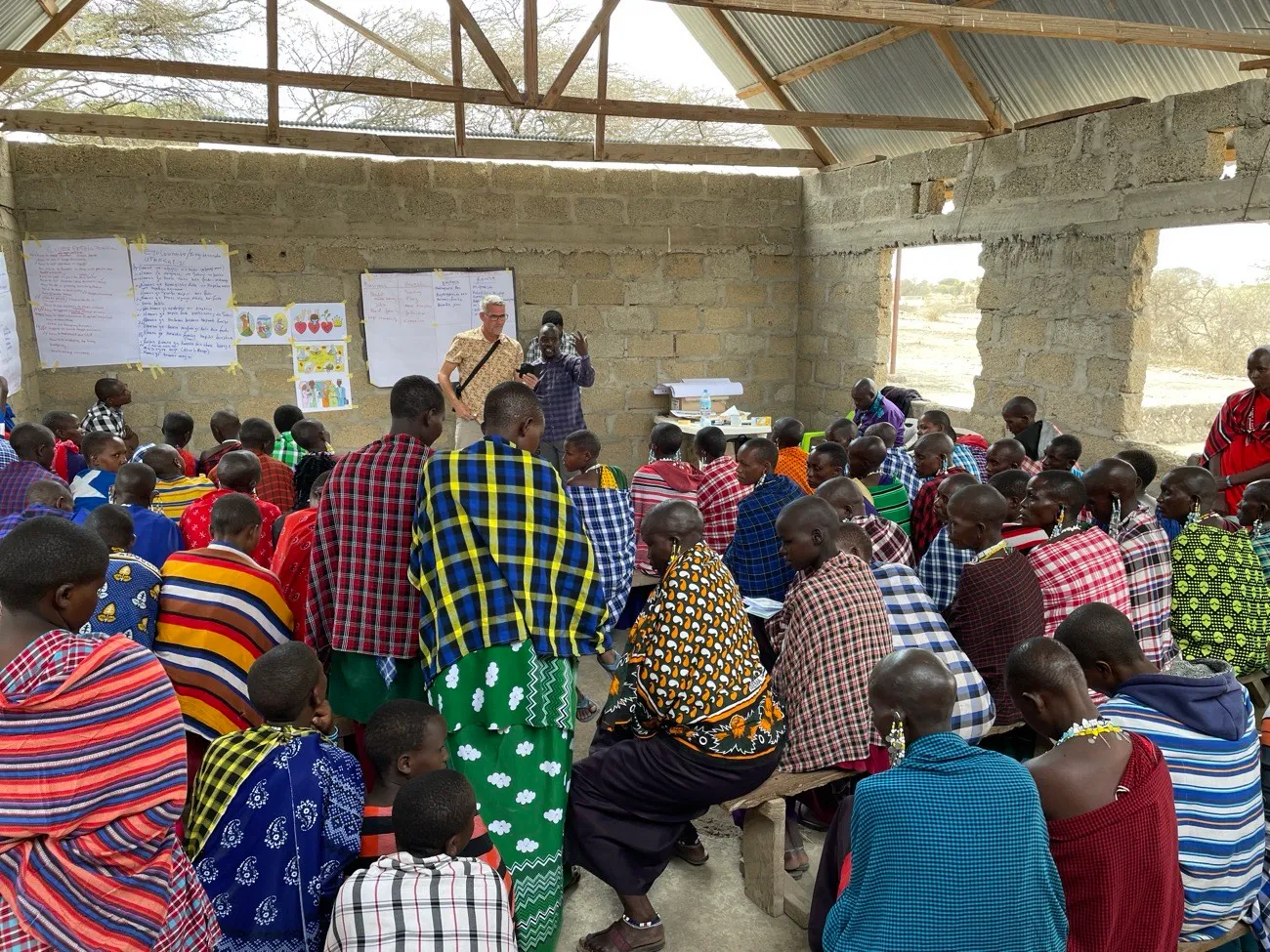 A large group in red and blue checked shukas sits on benches in an open-roofed building, listening to two trainers in front of handwritten flip charts, Tanzania.