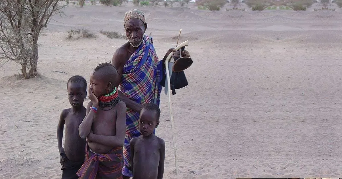 An elderly man in a checked blue and purple blanket, holding a walking stick, stands on dry sandy ground with three young boys.