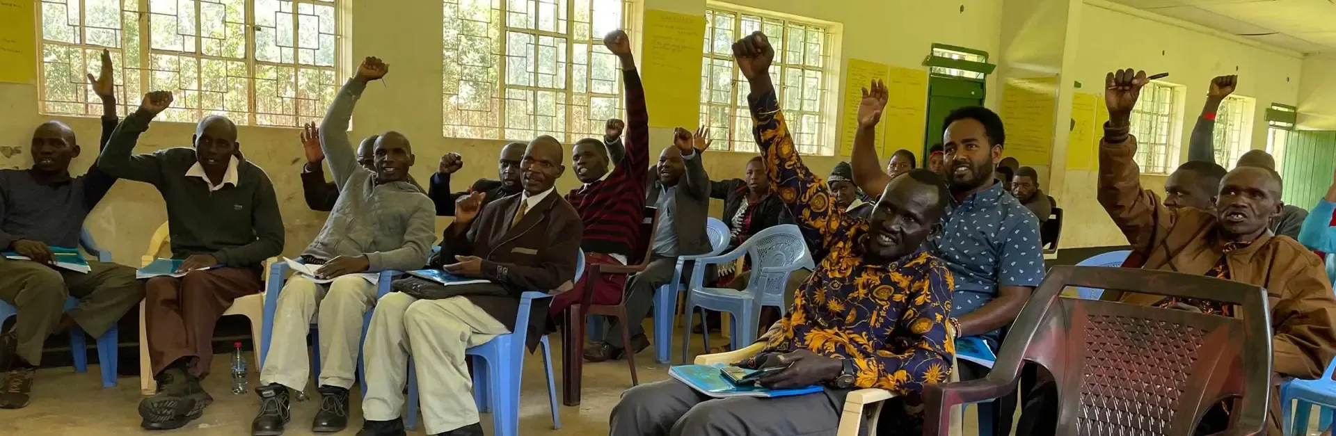 Training participants seated on plastic chairs raise their fists and hands in a bright classroom, Kenya.