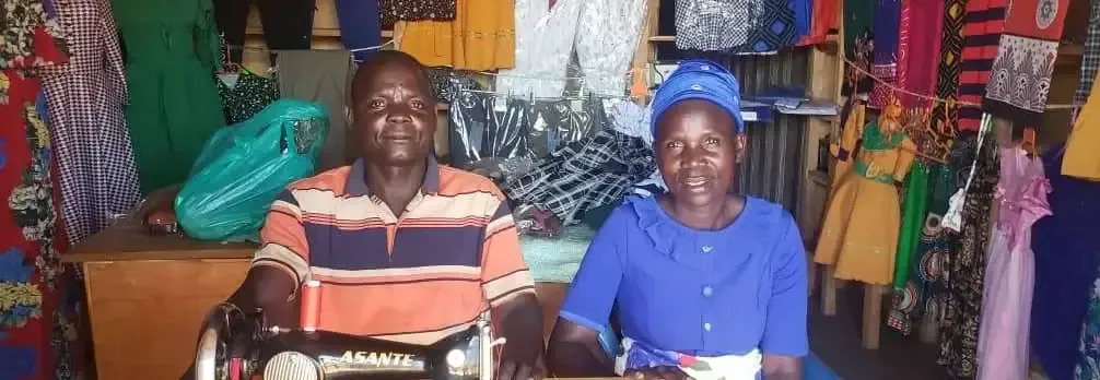 A man and a woman smile from behind a sewing machine in a small tailoring shop hung with colourful clothes and fabrics, Uganda.